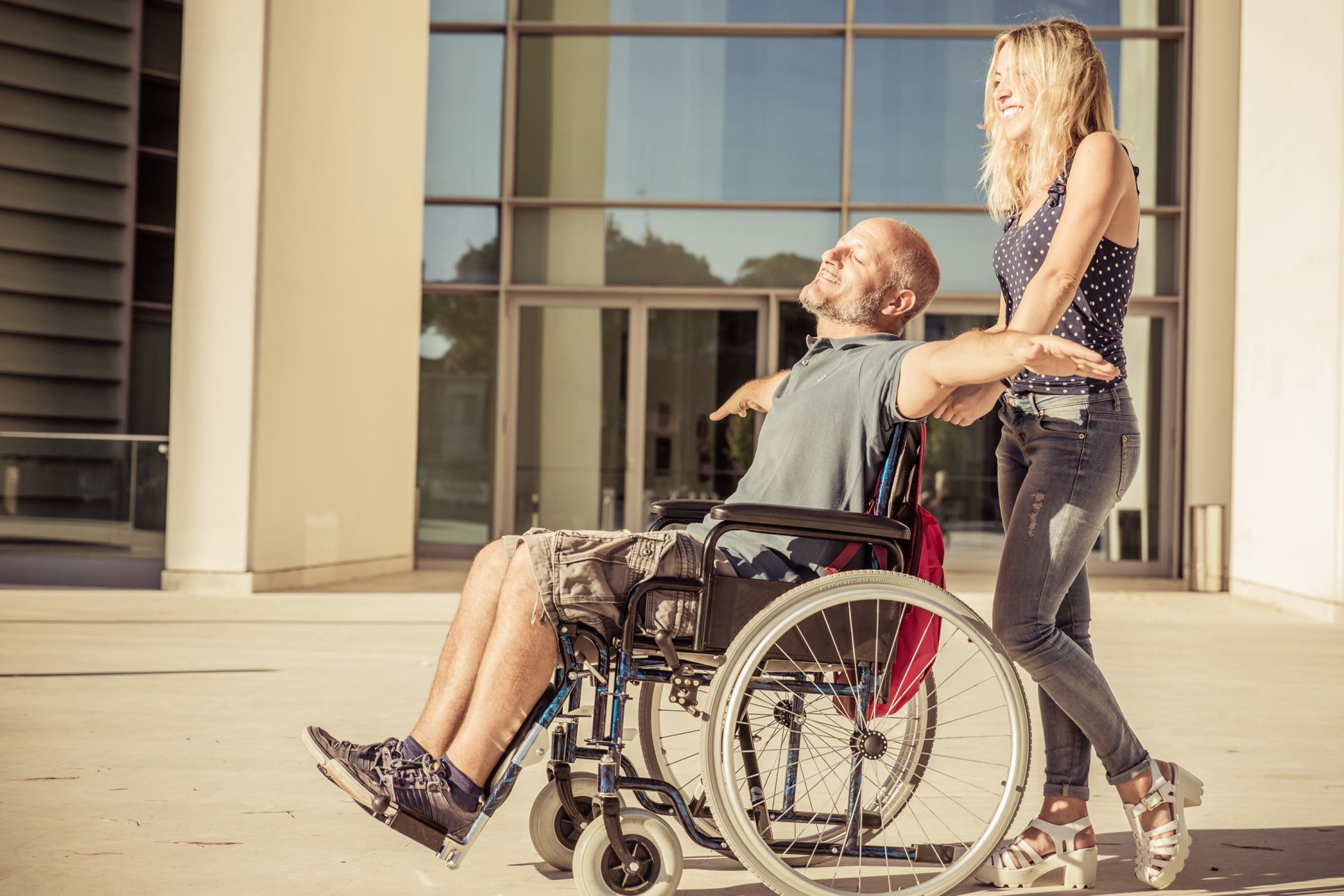 Image of a woman pushing a man in a wheelchair outside with his arms out like he is flying.