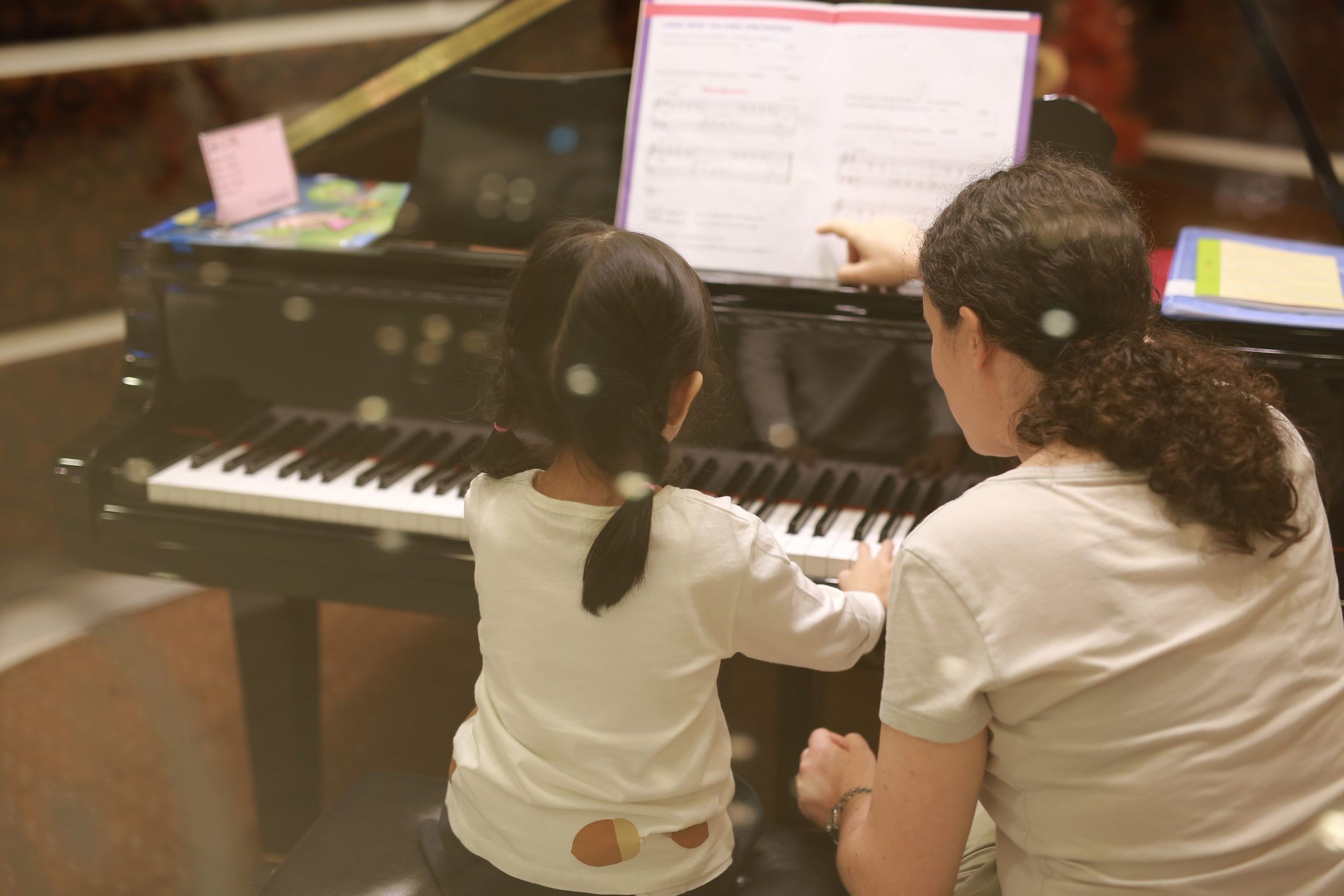 Little girl playing at a piano with her woman beside her pointing to the music.