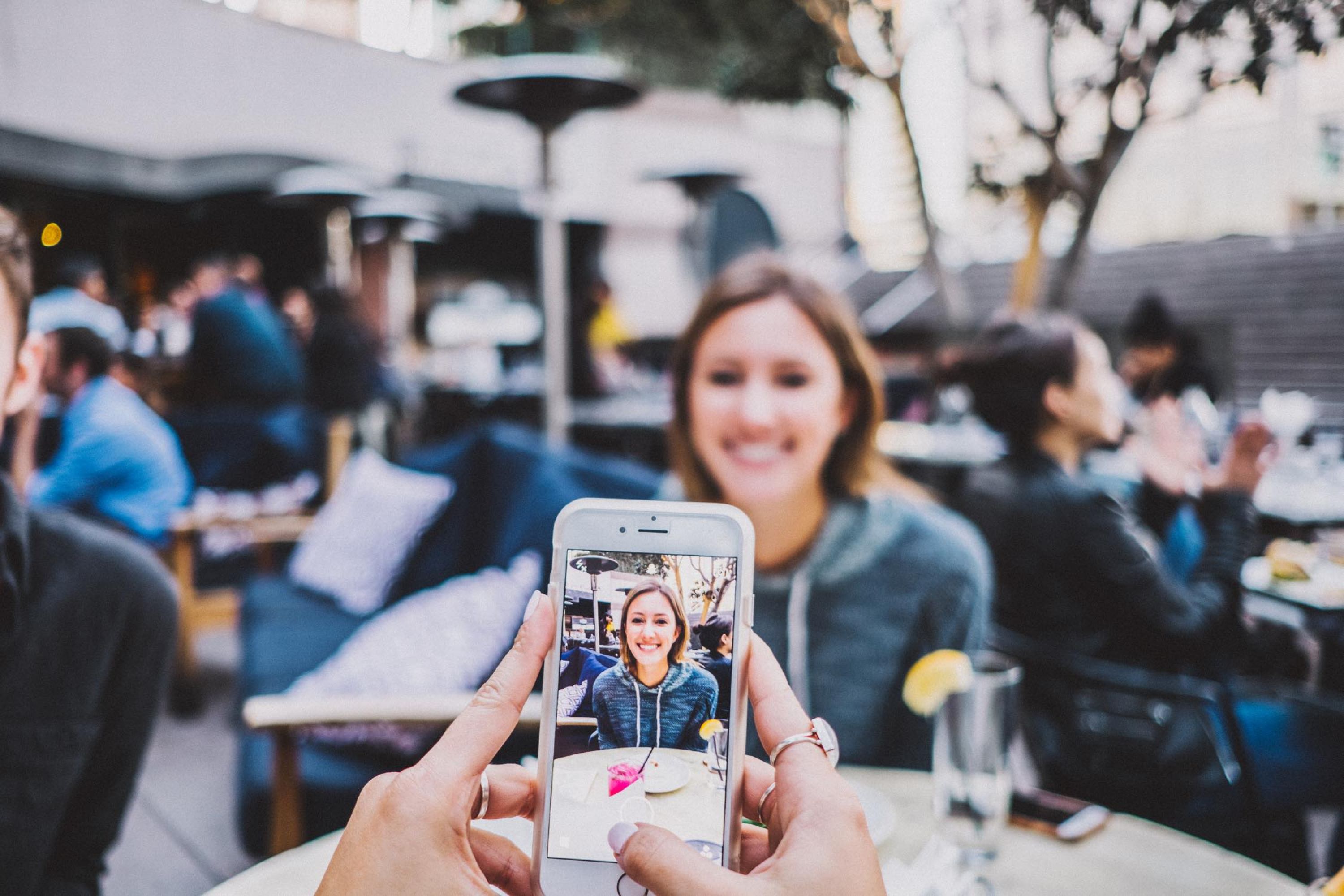 Image of hands holding an iPhone taking a photo of a girl sitting at a table on an outside patio.