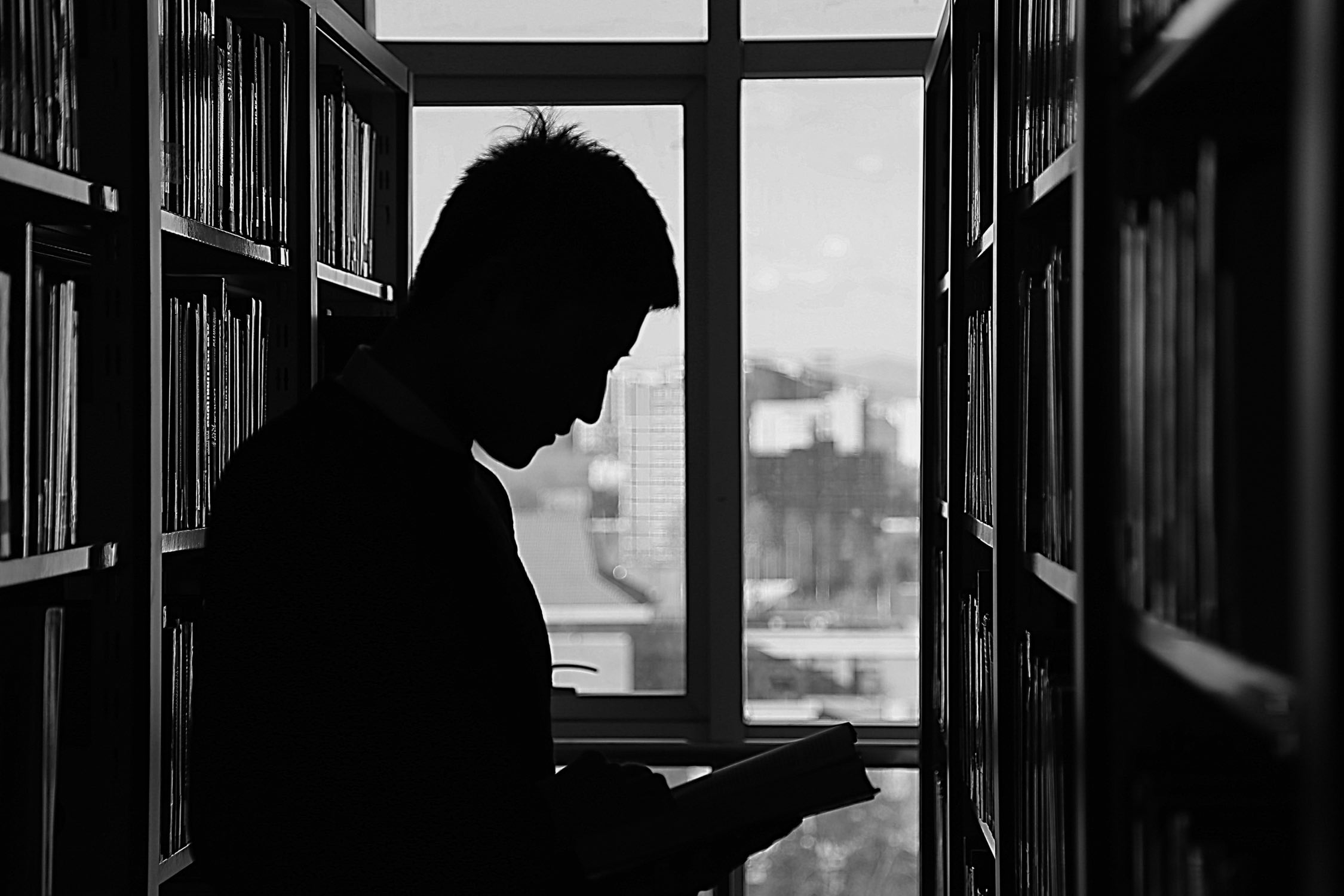 Black and white of a man's silhouette reading a book within a library.